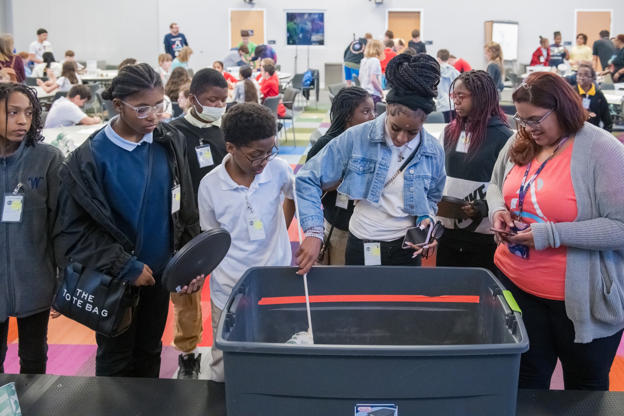 A group of students gather around a large, square container. One student moves items in the container with a stick. An adult at the right takes photos of the activity.