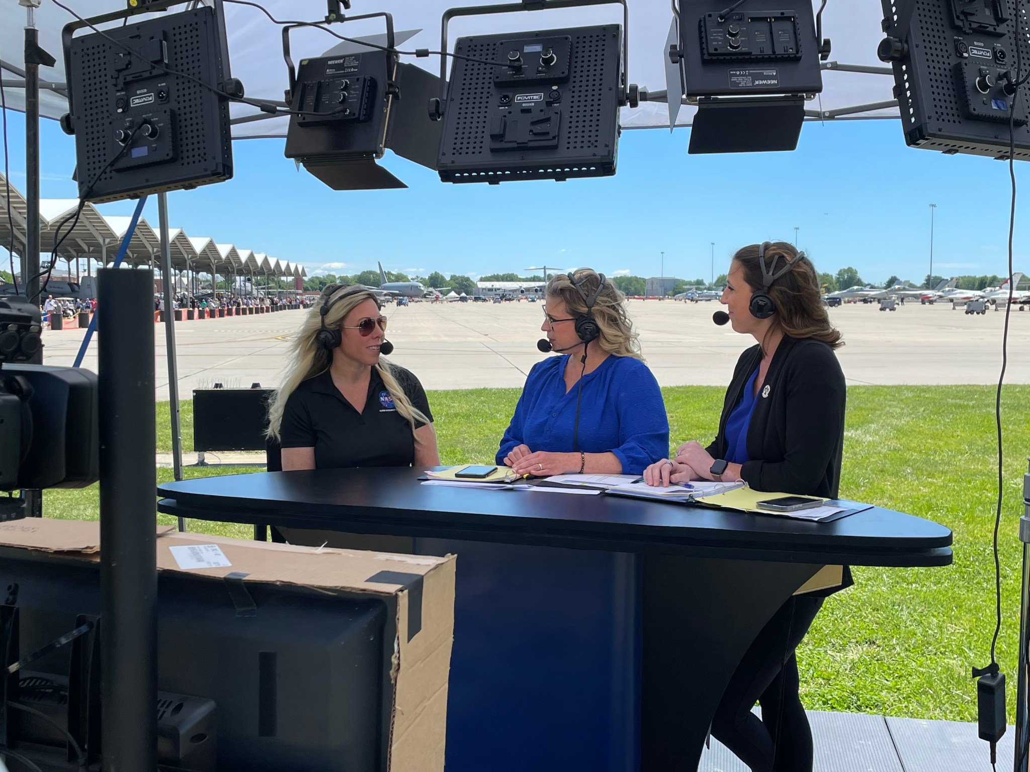 Three women interact as they sit on a stage in front of a news-type desk surrounded with computer screens and positioned toward the audience. Several airplanes park in the distance.