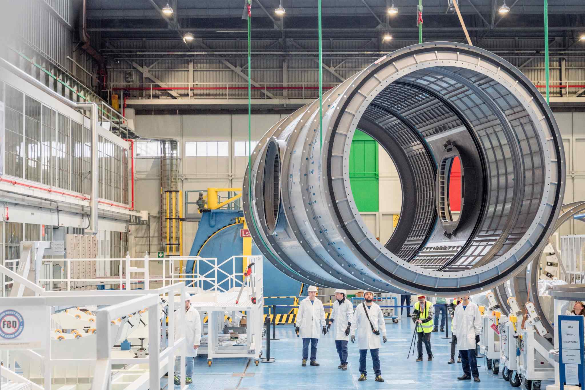 A large cylindrical module, the HALO (Habitation and Logistics Outpost) for NASA's Gateway space station, is being carefully moved inside a spacious industrial facility by technicians at Thales Alenia Space in Turin, Italy. The module is suspended by cranes and surrounded by workers in white lab coats and safety gear, preparing it for a series of stress tests.