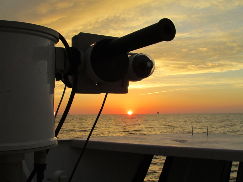 In the foreground of the image is a scientific instrument, which is shadowed due to the sunset behind it. In the background of the image, the ocean meets the sky at the horizon, and the sun is placed directly on the horizon emitting a yellow orange sunset that fades as it rises into the sky.
