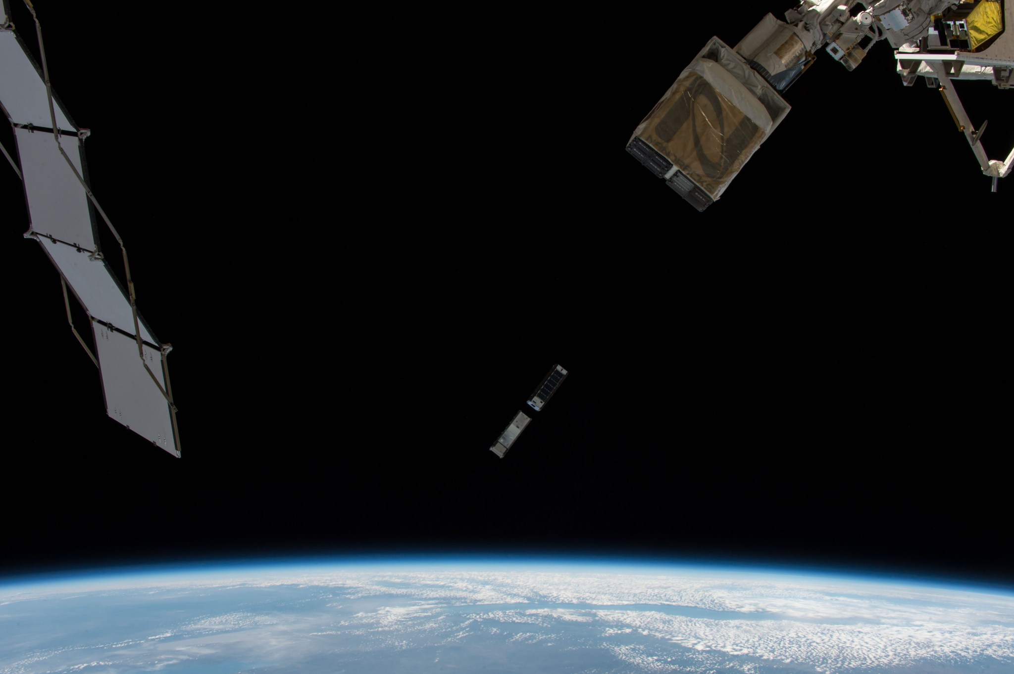 A blue, cloud-streaked Earth covers the bottom of this image. On the left is one of the station’s solar arrays and on the right, a large gold box that has just deployed two rectangular CubeSats, visible against the black background of space.