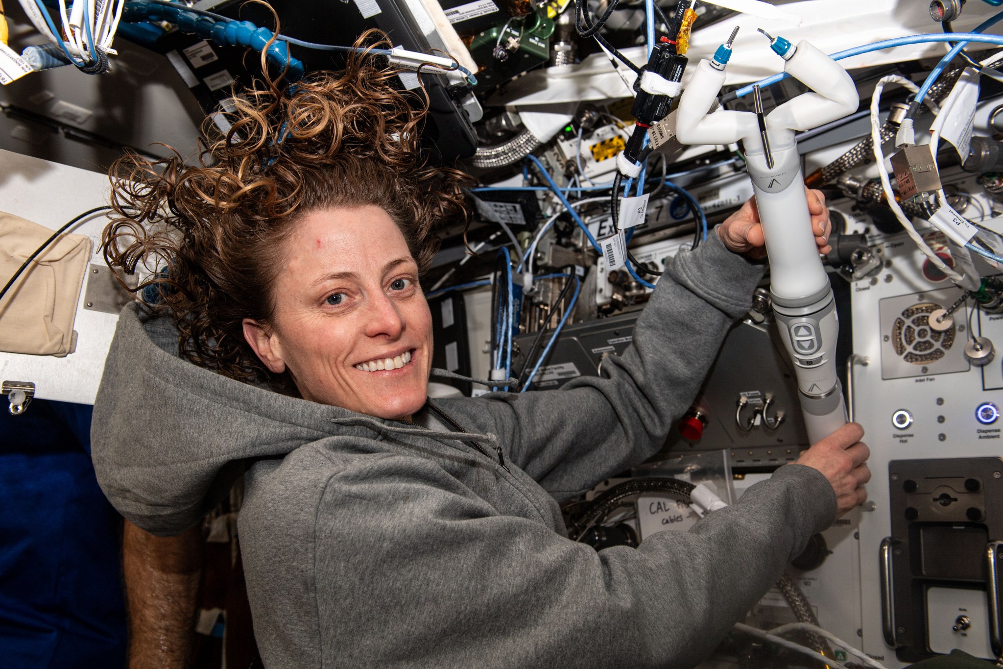 O’Hara is wearing a gray hooded sweatshirt and smiling at the camera, as she holds the miniature robot with both hands. The robot is narrow, about the size of an adult’s forearm, with a gray handle that has up and down arrow buttons and two bent “arms” on its end. One of the arms has a metal spatula and the other a set of clippers.