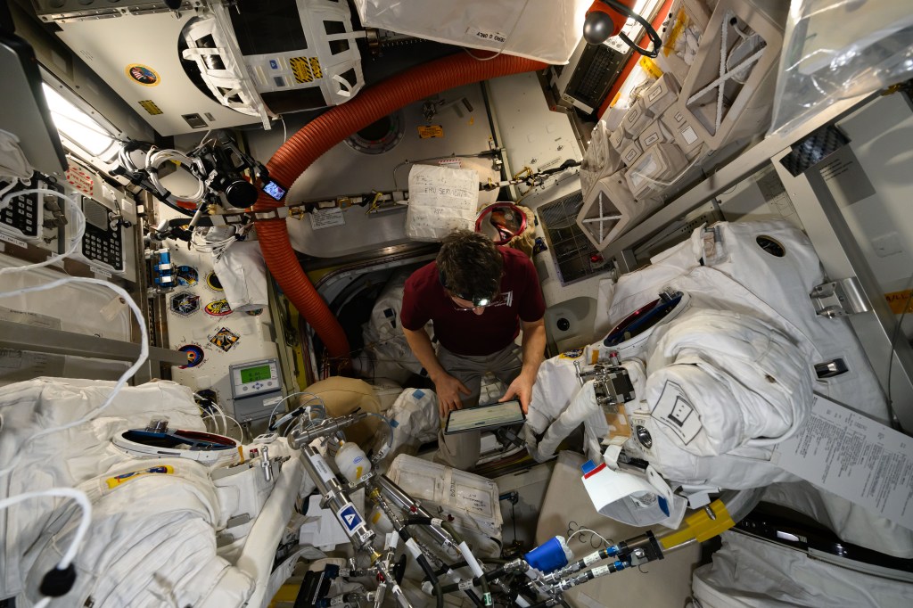 Expedition 71 Flight Engineer and NASA astronaut Mike Barratt reviews procedures on a computer tablet during maintenance on a pair of spacesuits inside the International Space Station's Quiest airlock.