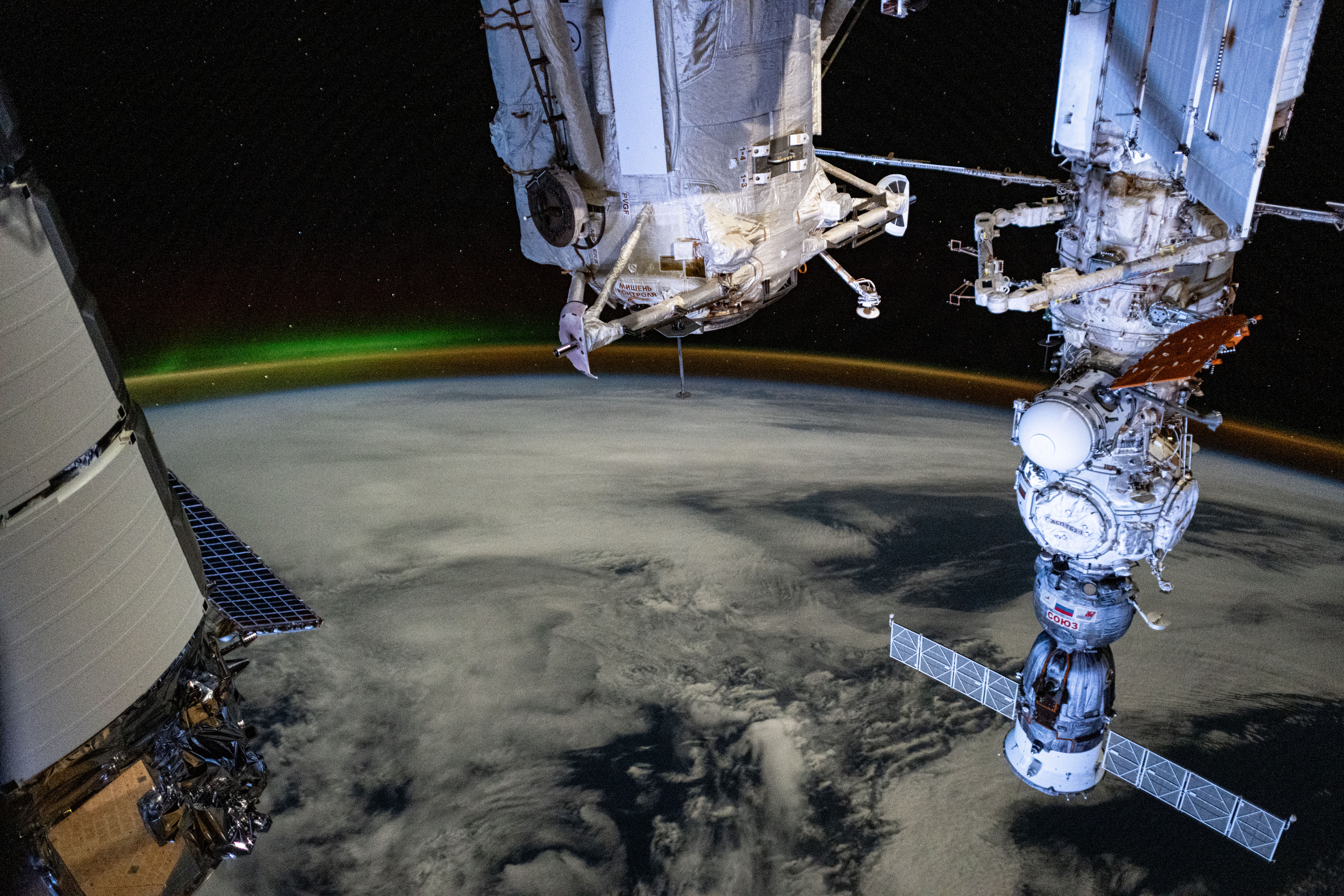 A dim aurora and an atmospheric glow crown Earth's horizon in this photograph from the International Space Station as it orbited 266 miles above a cloudy Indian Ocean southwest of Australia. In the foreground, from left, are a portion of Northrop Grumman's Cygnus space freighter, the Rassvet module, and the Soyuz MS-25 crew ship docked to the Prichal docking module which is itself mated to the Nauka science module.