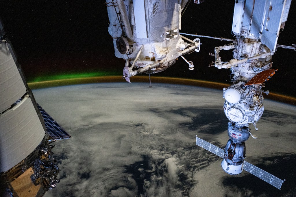 A dim aurora and an atmospheric glow crown Earth's horizon in this photograph from the International Space Station as it orbited 266 miles above a cloudy Indian Ocean southwest of Australia. In the foreground, from left, are a portion of Northrop Grumman's Cygnus space freighter, the Rassvet module, and the Soyuz MS-25 crew ship docked to the Prichal docking module which is itself mated to the Nauka science module.