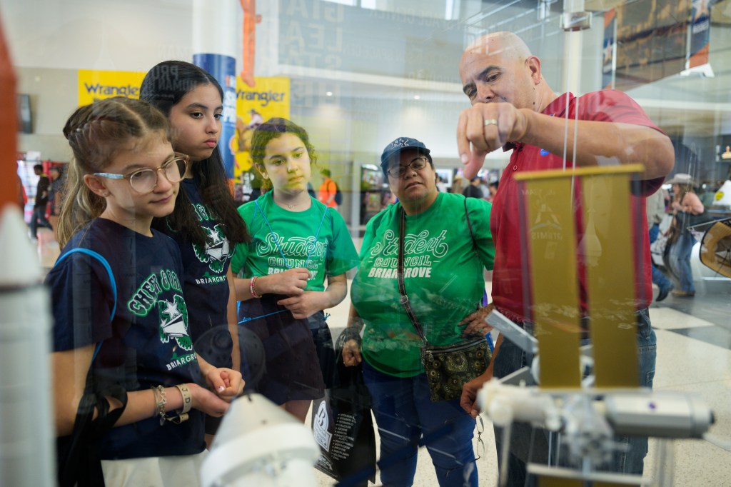 A NASA personnel member in a red shirt explains the Gateway space station model to three children and an adult. The group is gathered around the display, listening intently to the detailed explanation. The setting is a busy indoor venue