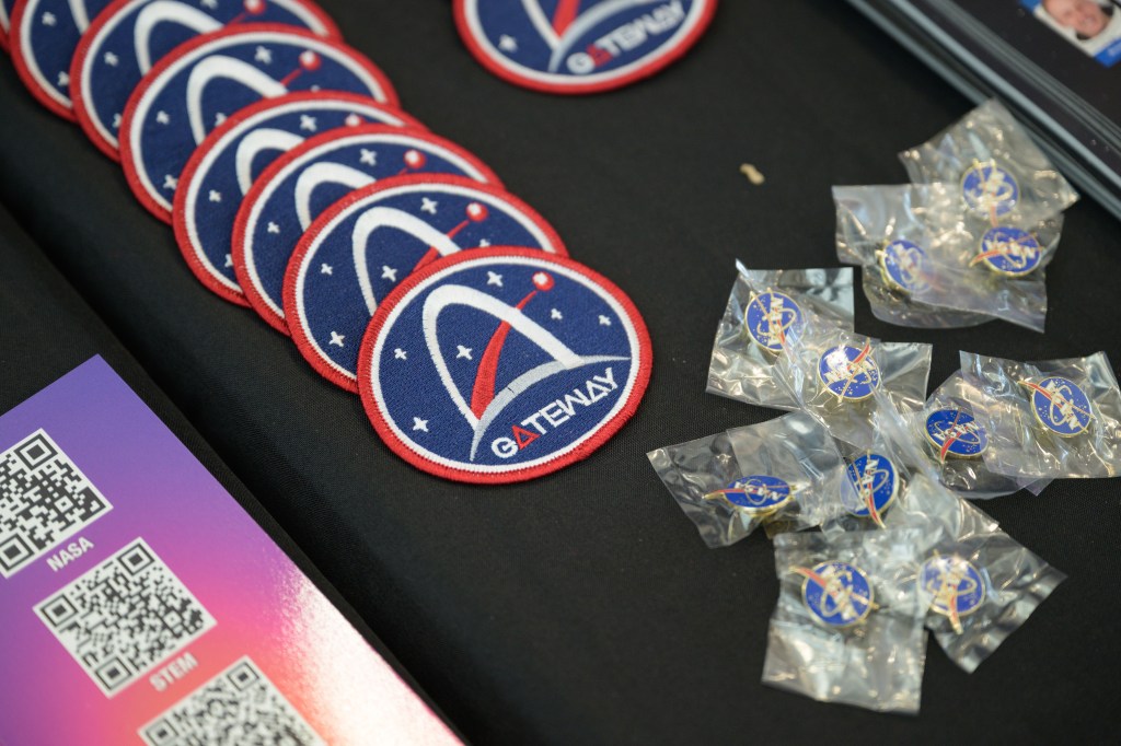 Close-up of Gateway patches and NASA pins neatly arranged on a black tablecloth at an outreach event. The patches feature the Gateway logo, and the pins are sealed in small plastic bags. A flyer with QR codes is also visible on the table, highlighting the available merchandise and promotional materials.