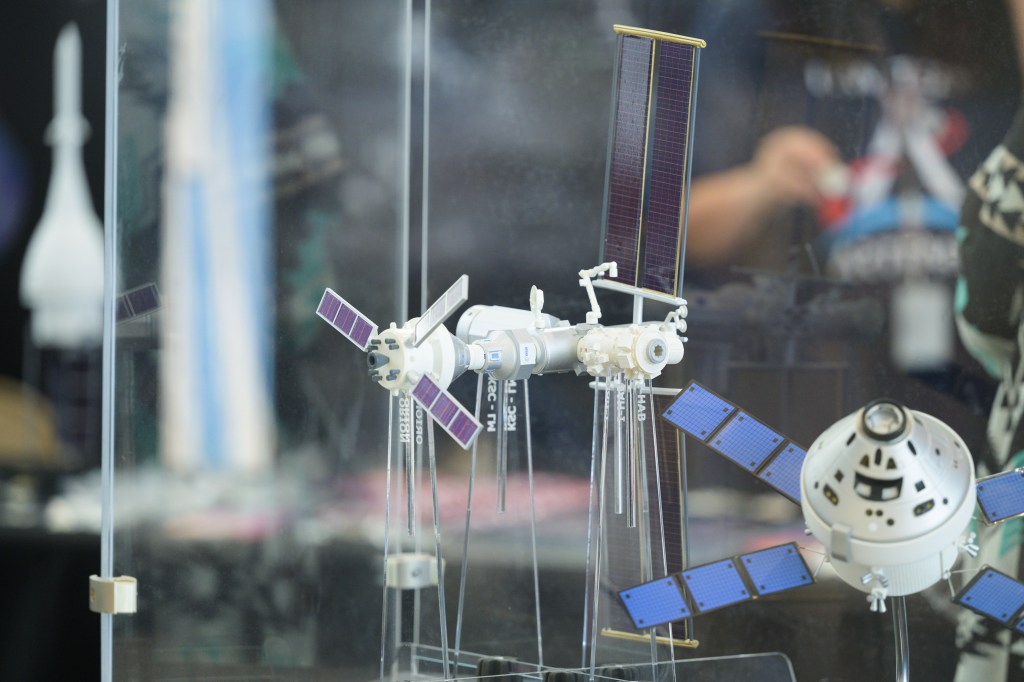 A close-up view of a detailed model of the Gateway space station, showcasing its various components and solar panels. The model is enclosed in a glass case, with a blurred background of the event venue.