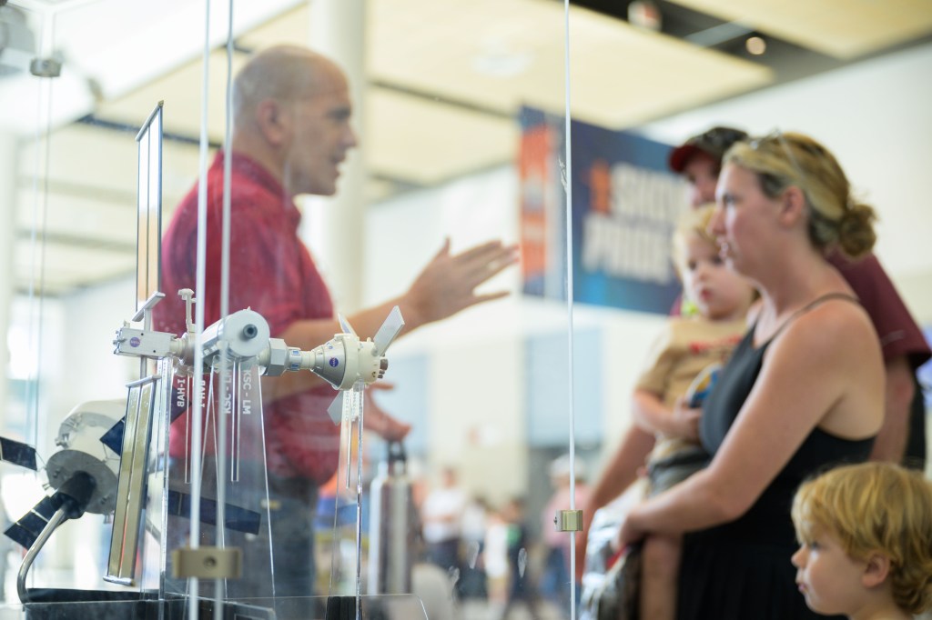A NASA personnel member in a red shirt engages with a family, including a woman holding a young child and another child standing beside her, at NASA's outreach event. The Gateway space station model is prominently displayed in the foreground. The scene is set in a bright, indoor venue.