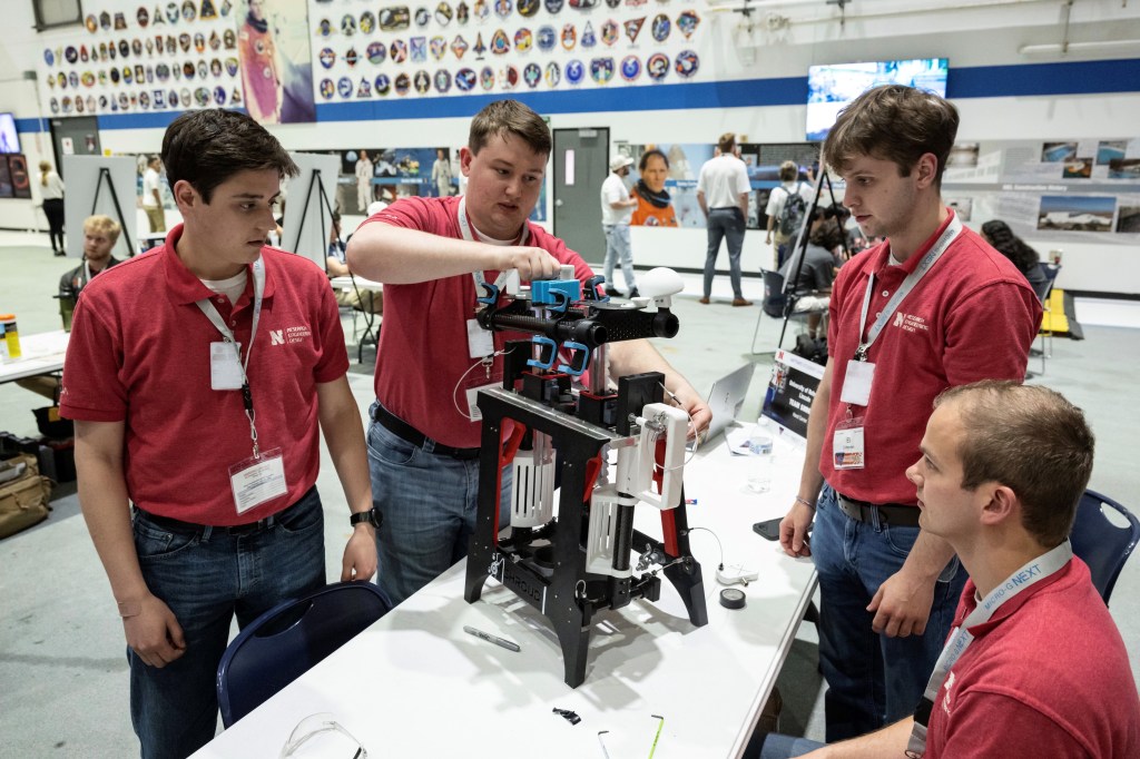 Four students in red shirts work on creating a tool on a white table.
