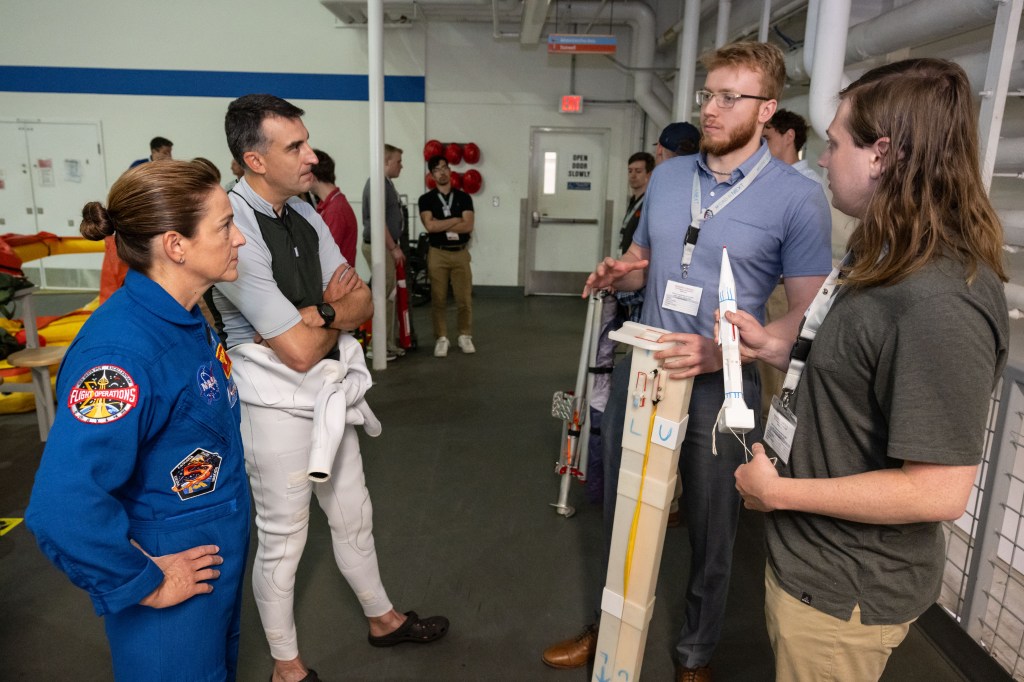 An astronaut in a blue flight suit and a diver talks to two students holding a lunar flagpole prototype.