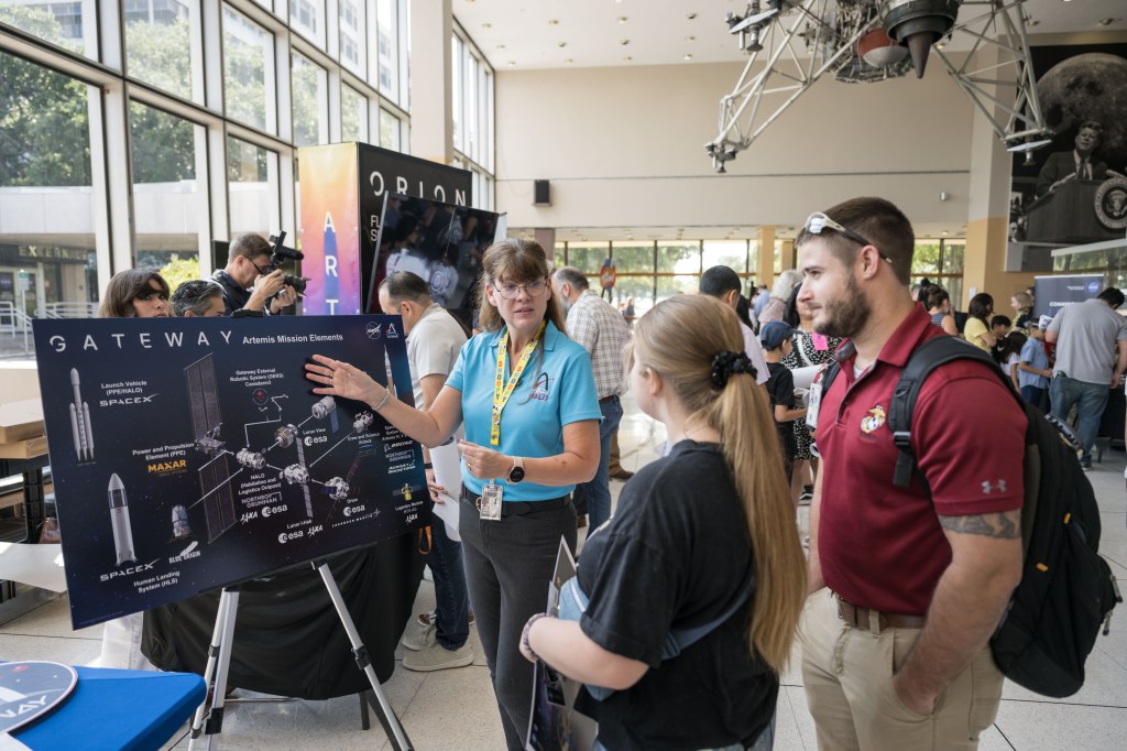 A Gateway Program employee in a blue shirt explains the Artemis Mission Elements using a large informational poster to a man and a woman at an event. The poster includes detailed diagrams of the Gateway space station, launch vehicles, and various mission components. The employee gestures towards different parts of the poster, engaging the visitors in discussion. The event is bustling with activity, with other booths and attendees visible in the background. The indoor venue features large windows that allow natural light to fill the space, creating a bright and welcoming atmosphere.
