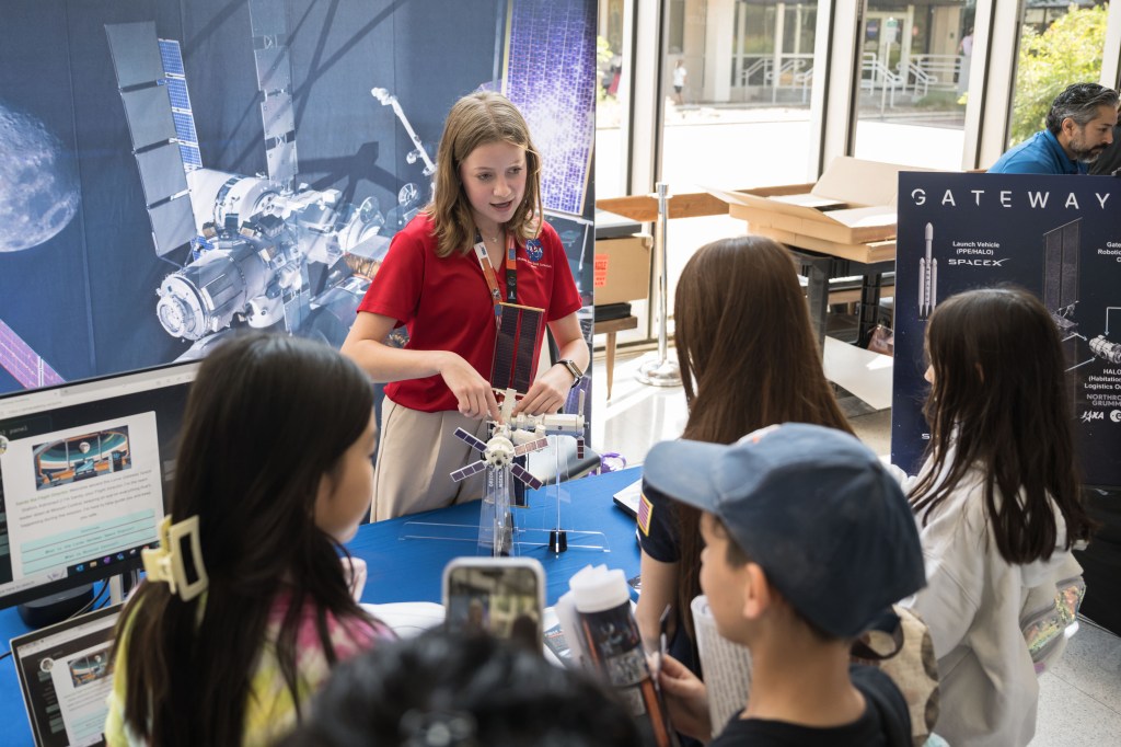 An intern in a red NASA shirt demonstrates a detailed model of the Gateway space station to a group of attentive children. The intern is explaining the features of the model, which includes solar panels and various modules. Behind the intern, a large poster of the Gateway space station and other informational displays are visible. The children are gathered around, some holding brochures, interested in the presentation. The event is taking place indoors, with natural light streaming in through large windows.