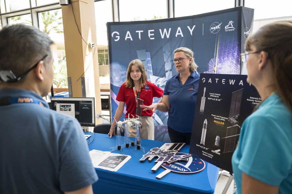 A young intern in a red NASA shirt and a Gateway Program employee in a blue shirt engage with visitors at a booth showcasing a model of the Gateway space station. The employee is explaining the model's features, while the intern listens attentively. Behind them, a large Gateway poster and other informational displays provide additional context. The booth includes detailed diagrams and models, highlighting various aspects of the Gateway space station.