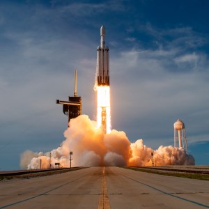 Looking down a runway, we see clouds of vapor billowing outward as a rocket takes off. A black tower structure called the gantry is on the left of the rocket's flames, and further in the distance on the right is a white water tower.