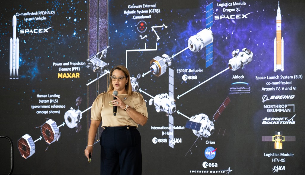 Dr. Dionne Hernandez-Lugo delivers a presentation on stage at the Shell Eco-marathon Americas event at the Indianapolis Motor Speedway on April 6, 2024. She is holding a microphone and gesturing while speaking about the Artemis Program. The large screen behind her shows detailed diagrams of the Gateway space station, various mission components, and partner organizations involved in the project.
