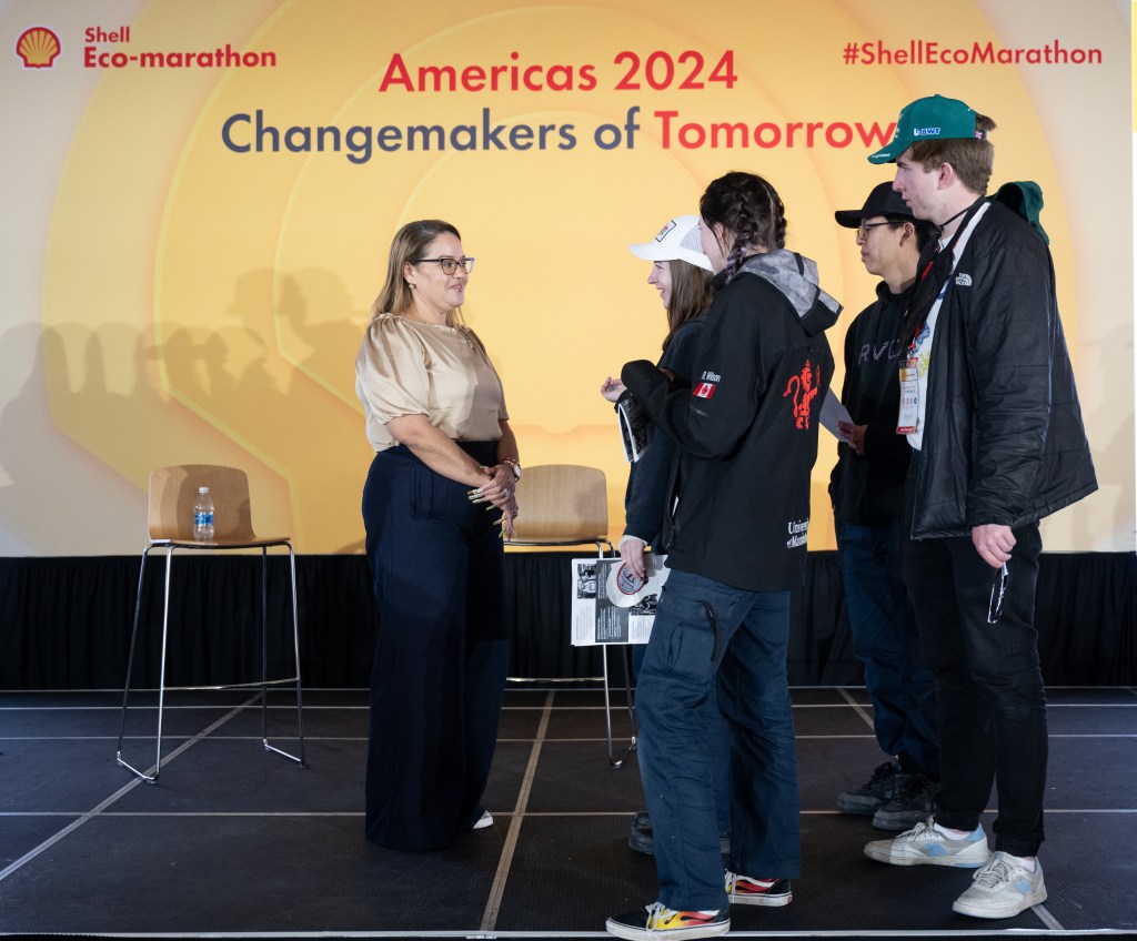 Dr. Dionne Hernandez-Lugo speaks with a group of students at the Shell Eco-marathon Americas event at the Indianapolis Motor Speedway on April 6, 2024. She is standing on a stage with two empty chairs and a table with a water bottle behind her. The students are listening attentively, holding pamphlets and engaging in conversation. The backdrop displays the event's theme, "Changemakers of Tomorrow."
