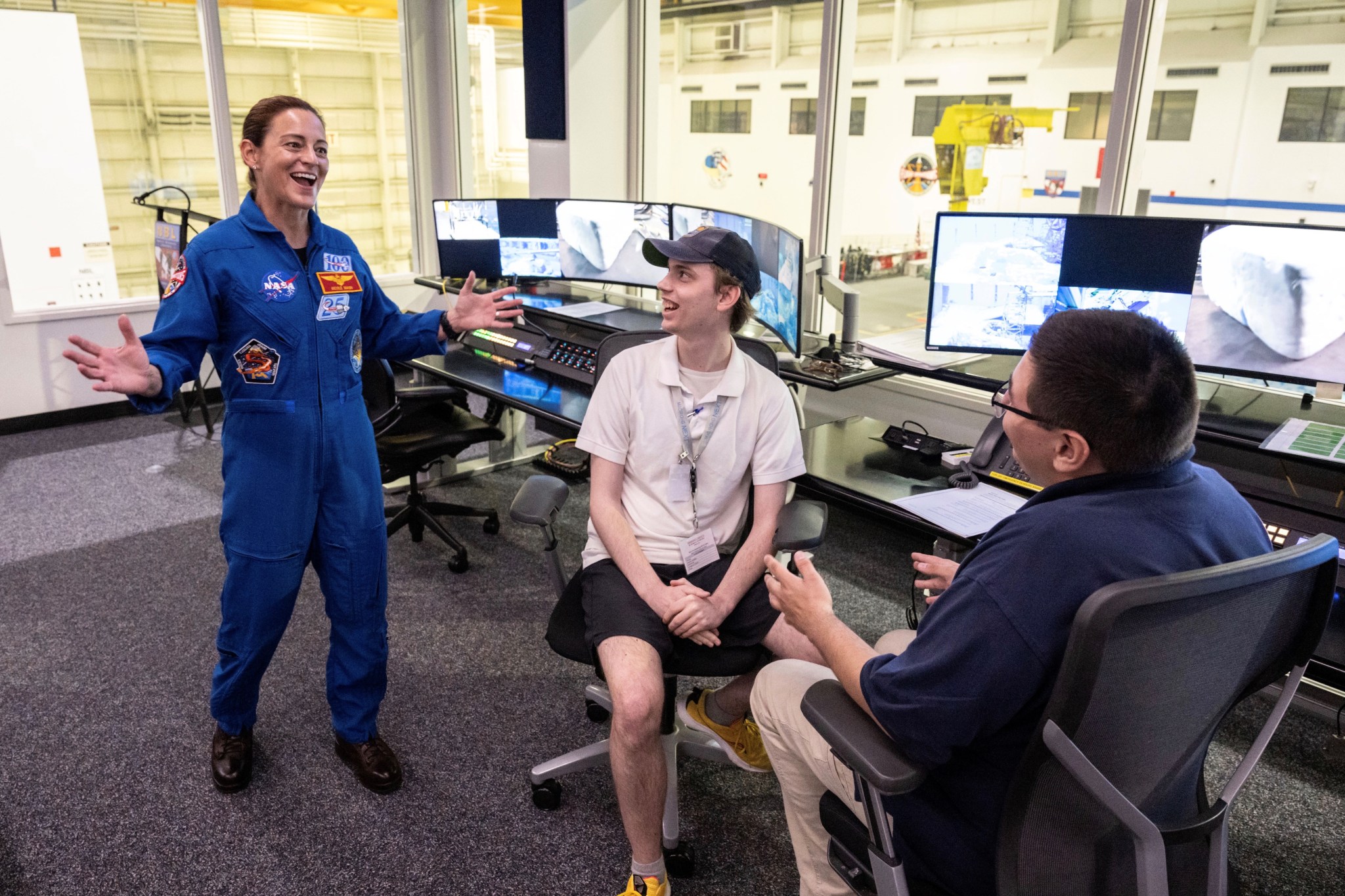 An astronaut wearing a blue flight suit laughts with students in a control room with screens showing underwater divers test their tools.