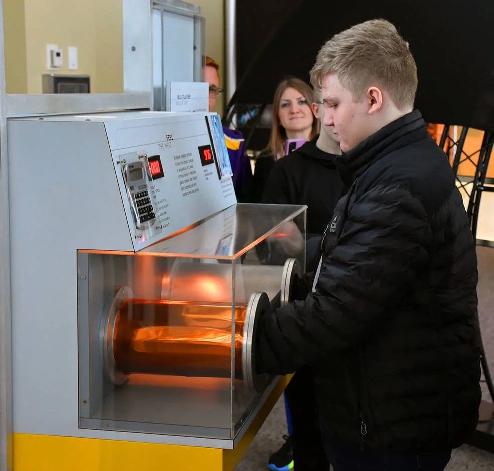 A visitor center guest stands with his hands in two tubes, one with thermal insulation and one without, to understand how temperature is controlled on the spacecraft.