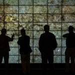 Four individuals stand in front of the NASA Advanced Supercomputing Facility hyperwall, a wall of LCD screens displaying an image of stars and galaxies.