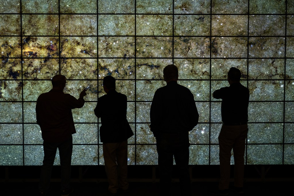 Four individuals stand in front of the NASA Advanced Supercomputing Facility hyperwall, a wall of LCD screens displaying an image of stars and galaxies.
