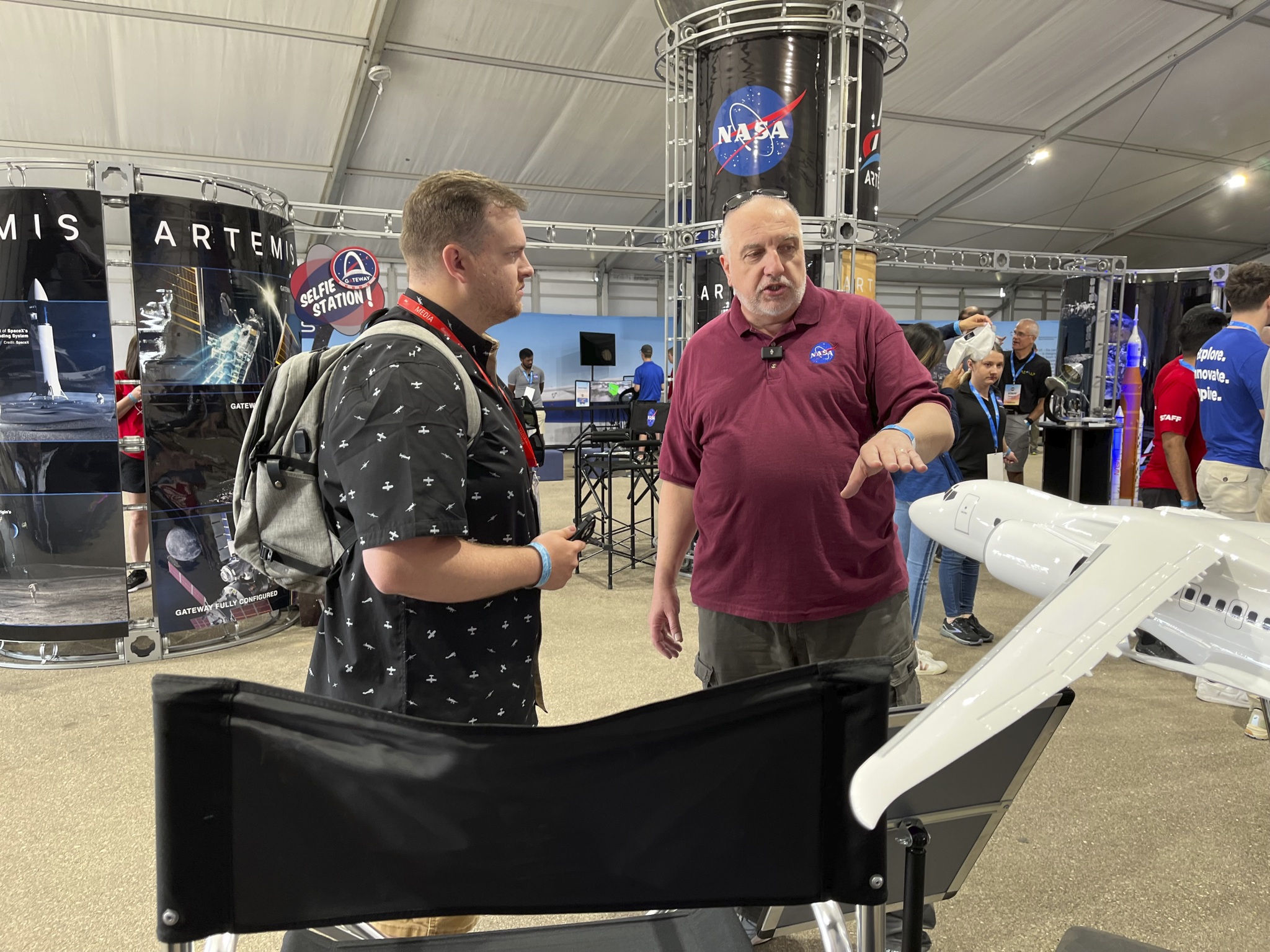 Two men stand in an exhibit tent next to a model of an airplane.