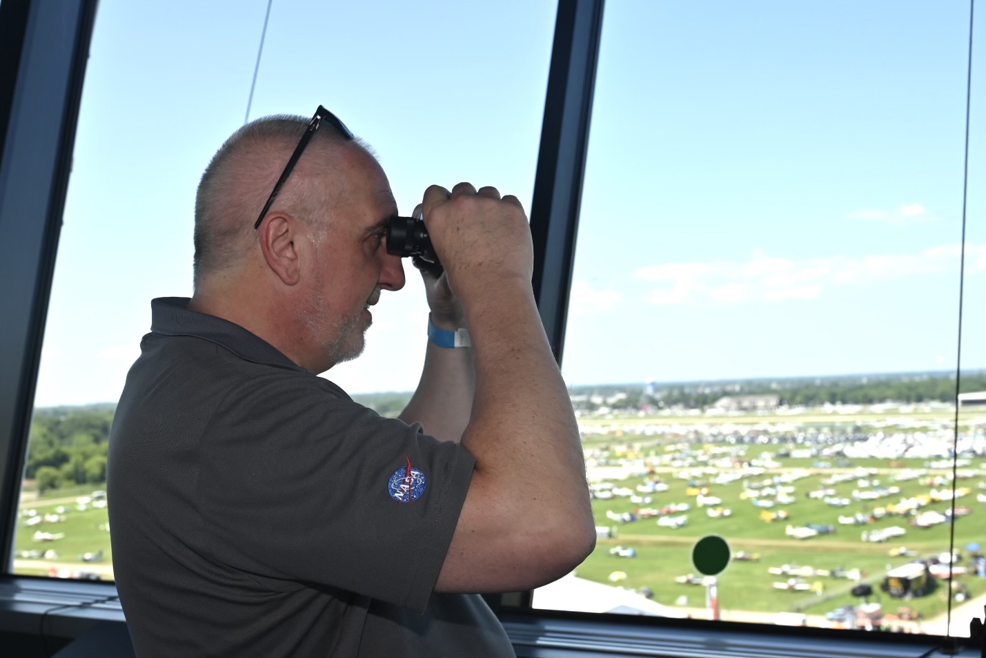 A man looks through a window using binoculars and sees hundreds of parked airplanes on the grass below.