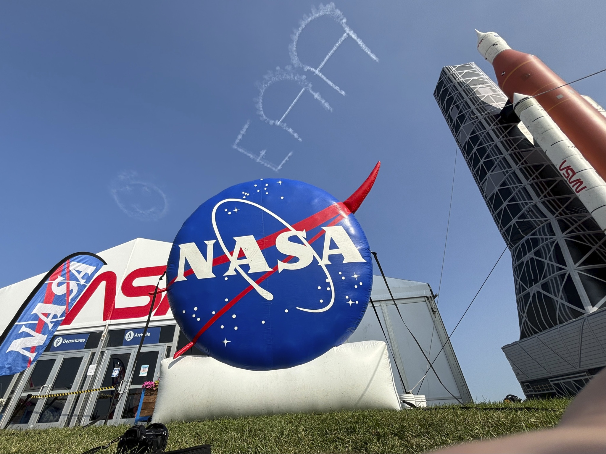 A giant inflatable NASA logo and rocket stand in a front of an exhibit tent beneath clear skies where a skywriting airplane has written EAA and a smiley face.