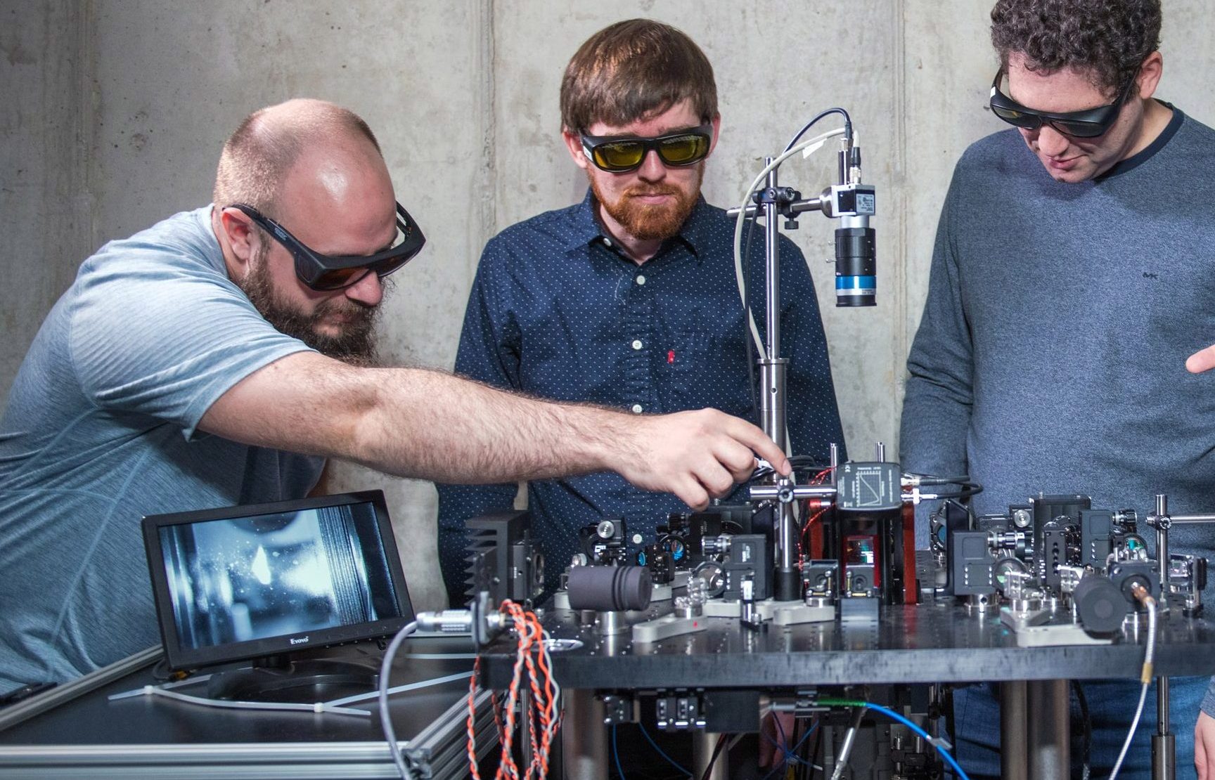 Five engineers inspect laboratory equipment. The quantum memory the equipment holds is shown on a digital screen. Engineers wear eye protection and point at the equipment.