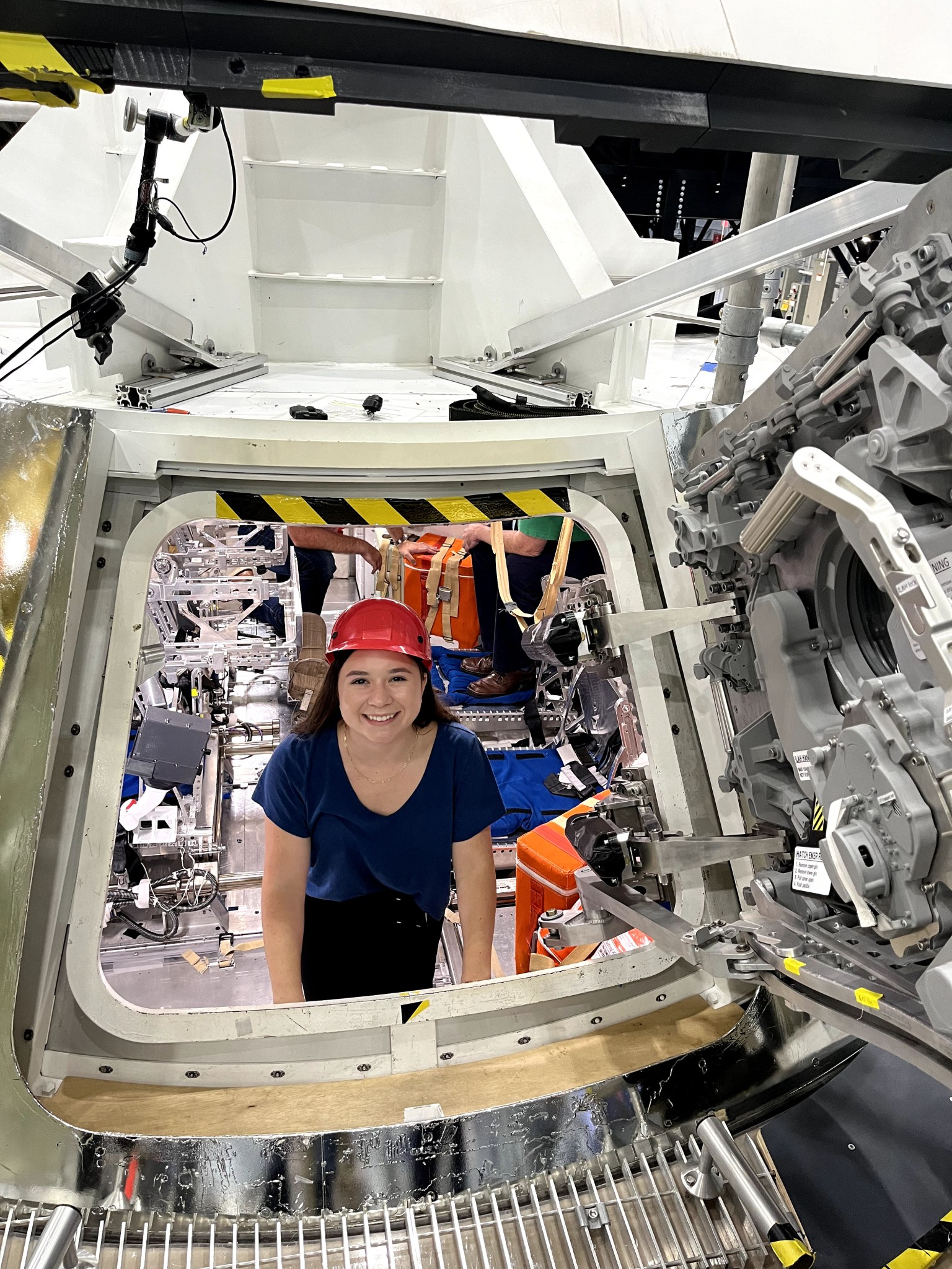 A young woman wearing a safety helmet poses inside of an Orion spacecraft mockup.