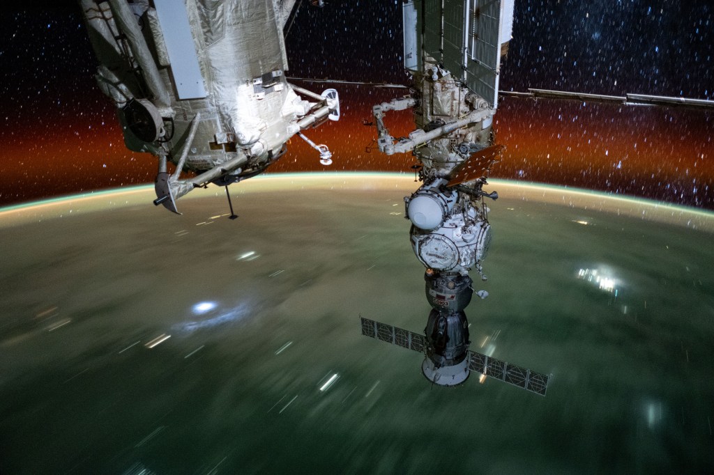 The long duration photograph from the International Space Station highlights the Rassvet module (left) and the Soyuz MS-25 spacecraft (right) docked to the Prichal module which is itself attached to the Nauka science module. 255 miles below the orbital outpost is a cloudy Pacific Ocean blanketing islands northeast of Indonesia's province of Papua. Above Earth's horzon is the planet's atmospheric glow and star trails glittering in the vastness of space.