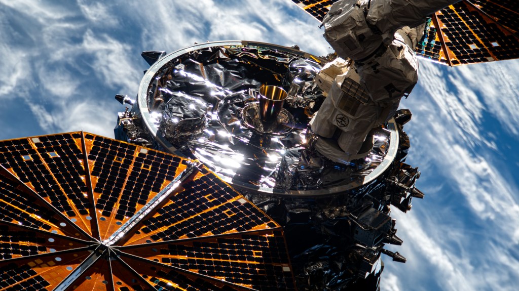 Northop Grumman’s Cygnus spacecraft is center of the image with two large gold solar panels, one in the top right and one in the bottom left corners. A large white metal arm is attached to the body of the spacecraft. Earth is visible in the background.