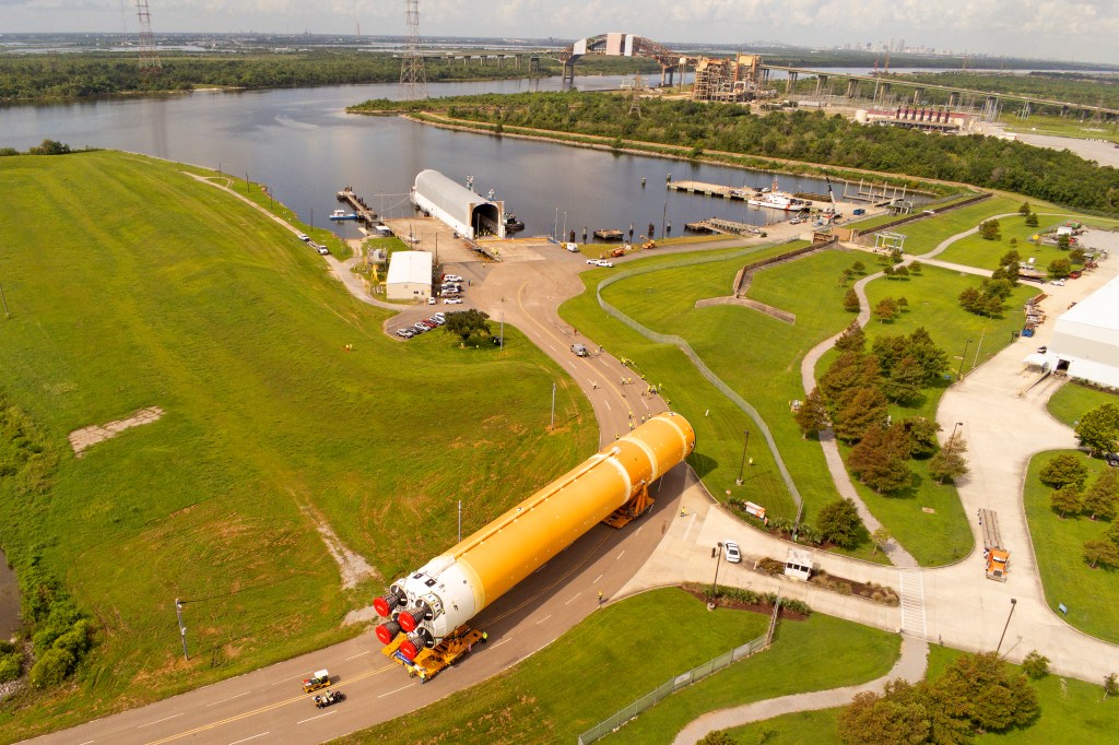 An aerial view of the Artemis II SLS core stage on a road. The core stage is a massive orange and white cylinder with four silver and red engines at the bottom. There are two vehicles on the road behind the core stage, highlighting its size. The road curves up from bottom left of the photo to the top middle, where it ends at a body of water. The land surrounding the road is a bright green.