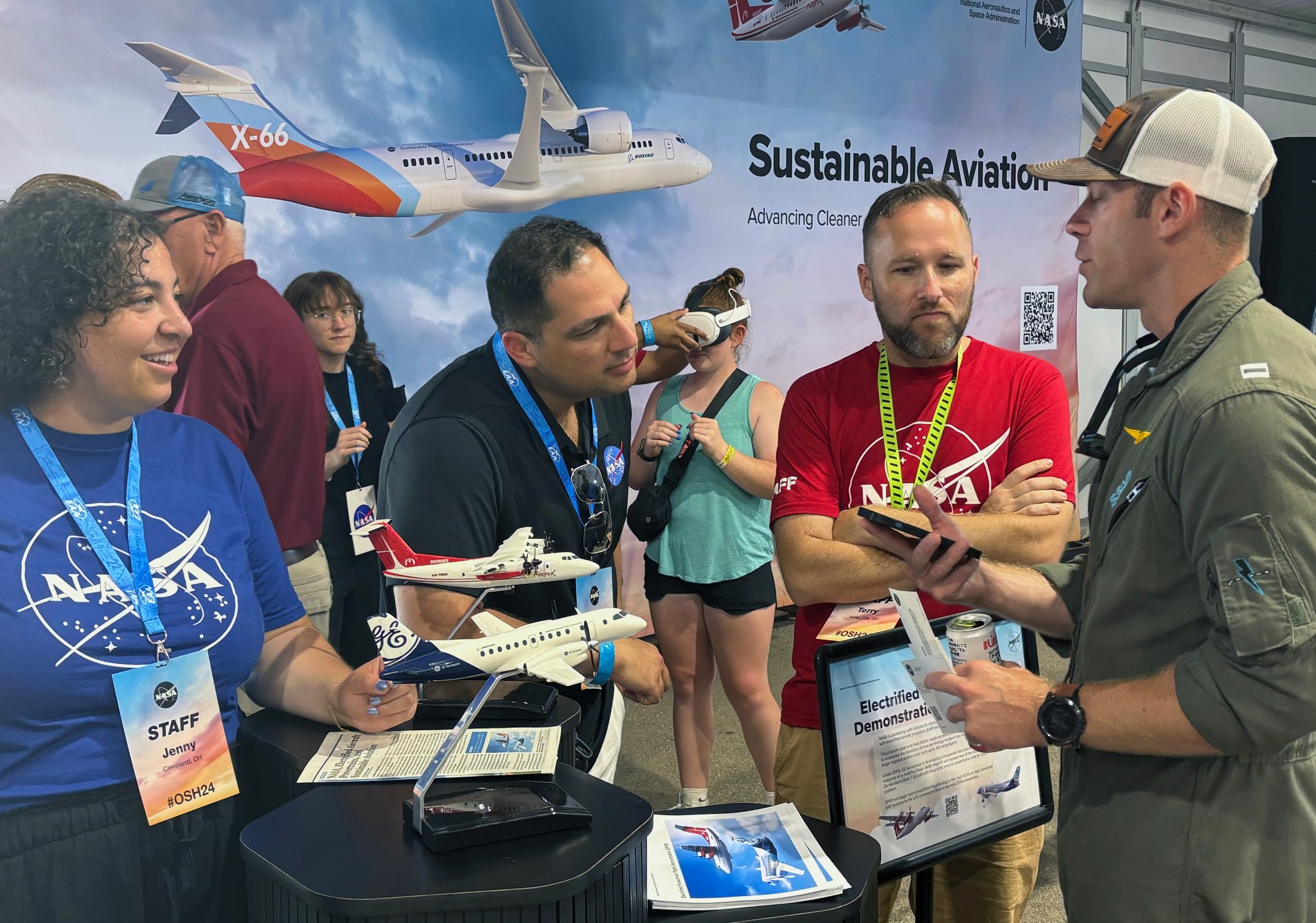 A group of people stand around a table that has two aircraft models and a stack of pictures on it.