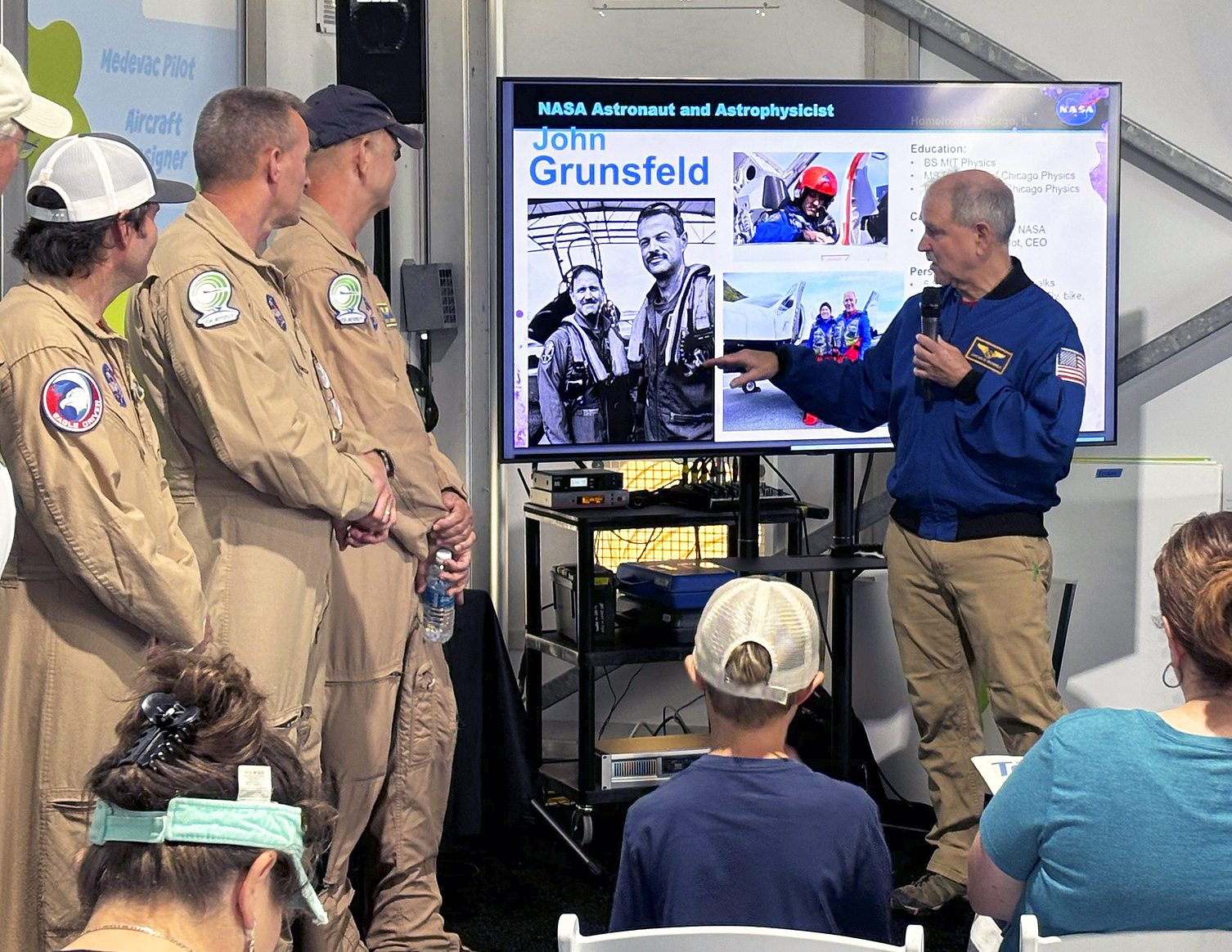 Three men in tan flight suits stand at left, Another man in a blue jacket stands at right pointing at a monitor displaying his biographical information. Audience members are seated in the foreground.