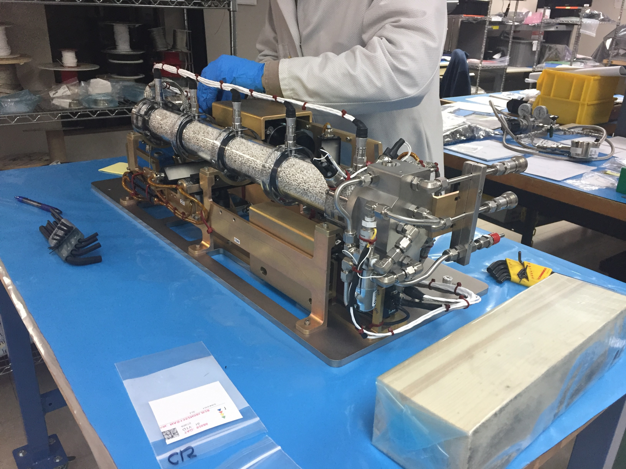 A suitcase-sized piece of equipment sitting on a blue tabletop has a copper-colored frame and a metal box on the closest end with multiple nozzles and cords. A clear tube the length of the hardware is filled with small white beads. A person wearing a white lab coat and blue gloves is visible from the shoulders down behind the equipment.