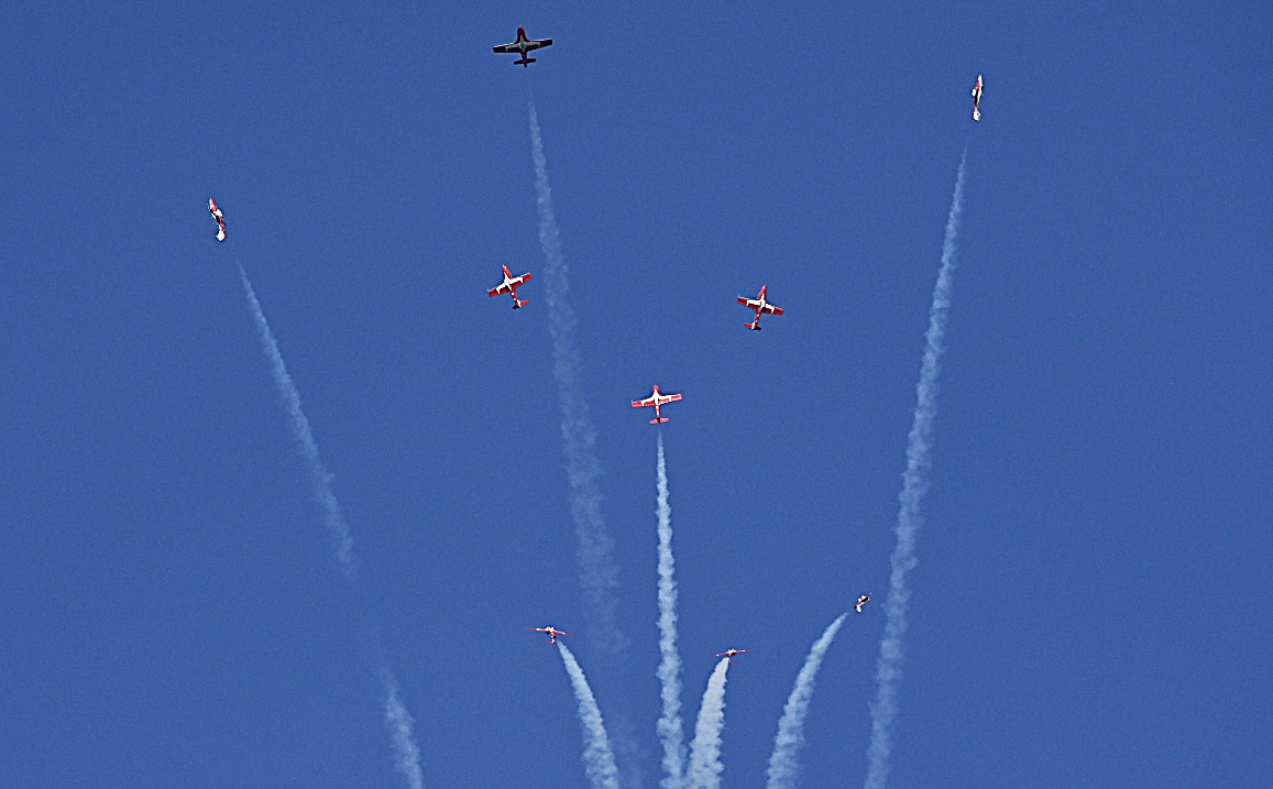 A group of nine jets with white smoke trailing behind them fly overhead in clear skies.