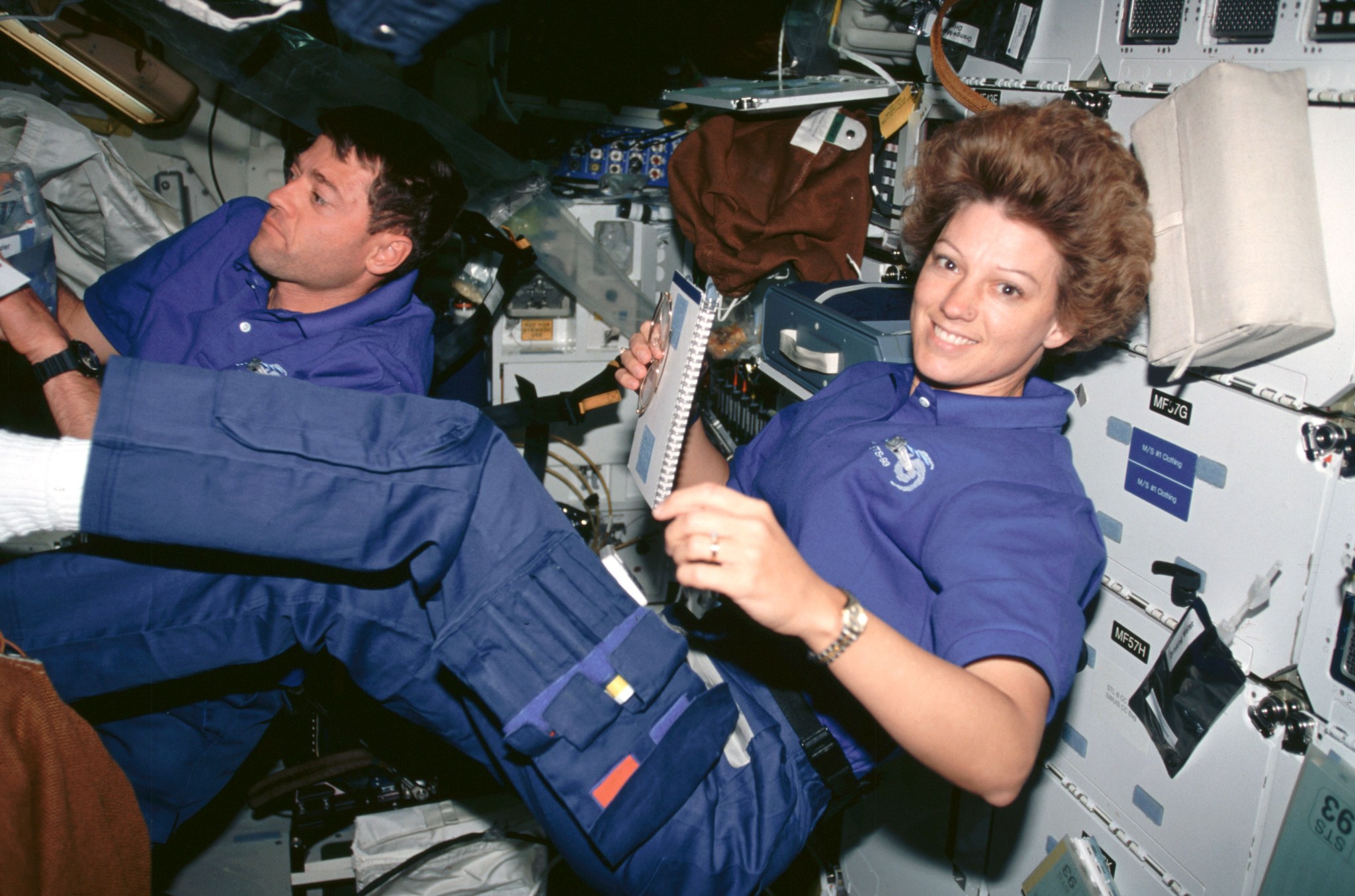 Two astronauts, one male (back) and one female (front) float inside the space shuttle. The woman in the front smiles at the camera while holding a spiral bound notebook in her right hand. She is wearing a blue polo shirt and blue pants with utility pockets on the thigh, as well as a gold watch on her left wrist. Her short brown hair floats upward.