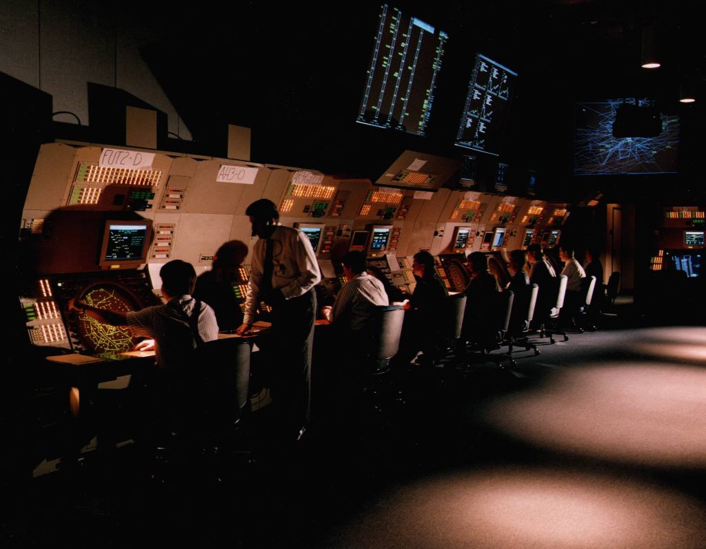 Several people sitting at a bank of radar screens in a darkened room. Three large screens overhead display different air traffic management tools.