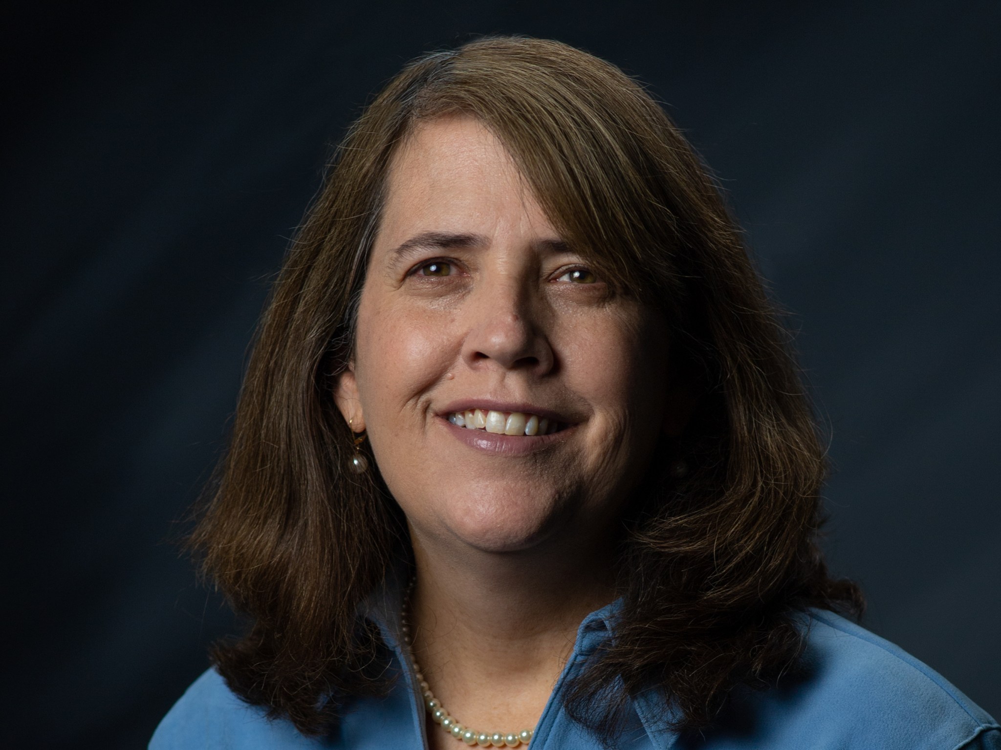 Center Deputy Director Christine Powell sits for a studio portrait wearing a blue collared shirt and a pearl necklace