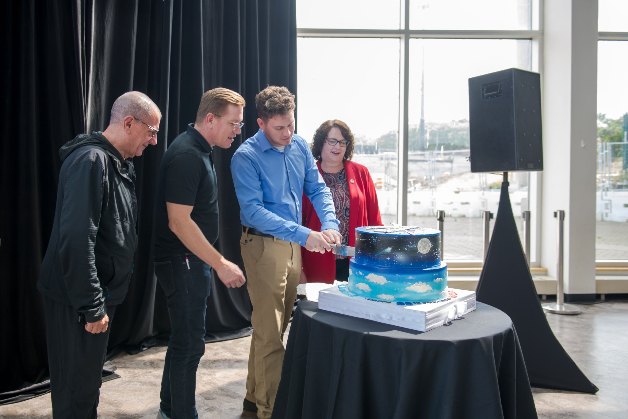A family member of the logo designer stands at left, while another cuts the cake that features clouds and planets. NASA Glenn’s Deputy Center Director stands on the right.
