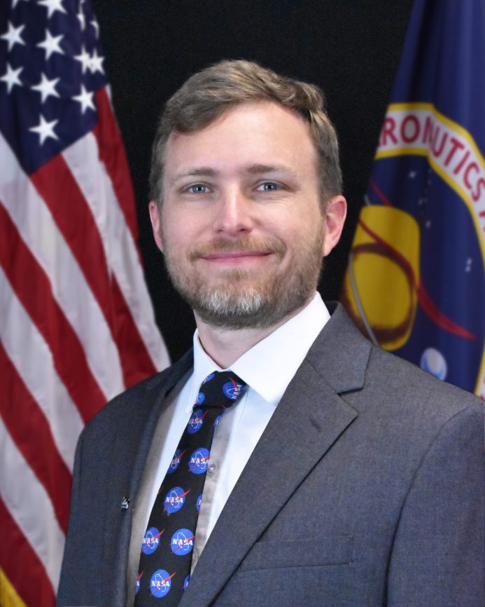 Headshot of Michael Kiskach, wearing a gray suit with a navy blue tie decorated with small NASA logos. He is smiling with his mouth closed. He is standing in front of an American flag and the NASA flag.