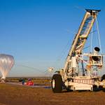 A crane holds a science instrument as a large scientific balloon, tethered to the ground, inflates before liftoff in New Mexico.