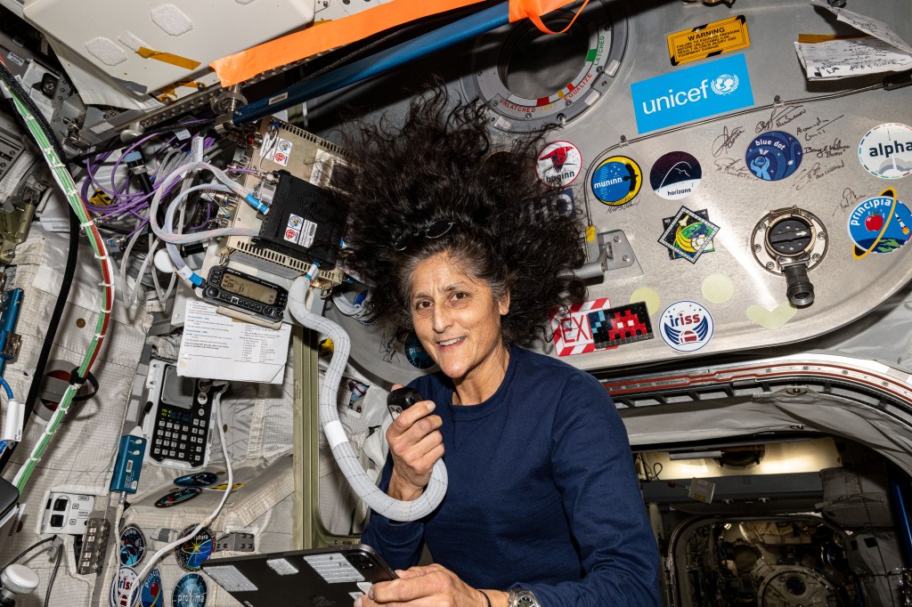 Williams, wearing a long-sleeved blue shirt, is facing the camera, and holding in her right hand a microphone attached by a large cord to a radio unit mounted on the wall. Her hair is floating around her head and the wall behind her is covered with various stickers.