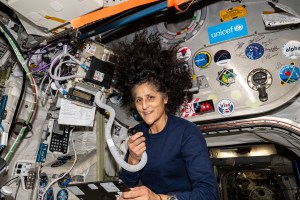 Williams, wearing a long-sleeved blue shirt, is facing the camera, and holding in her right hand a microphone attached by a large cord to a radio unit mounted on the wall. Her hair is floating around her head and the wall behind her is covered with various stickers.