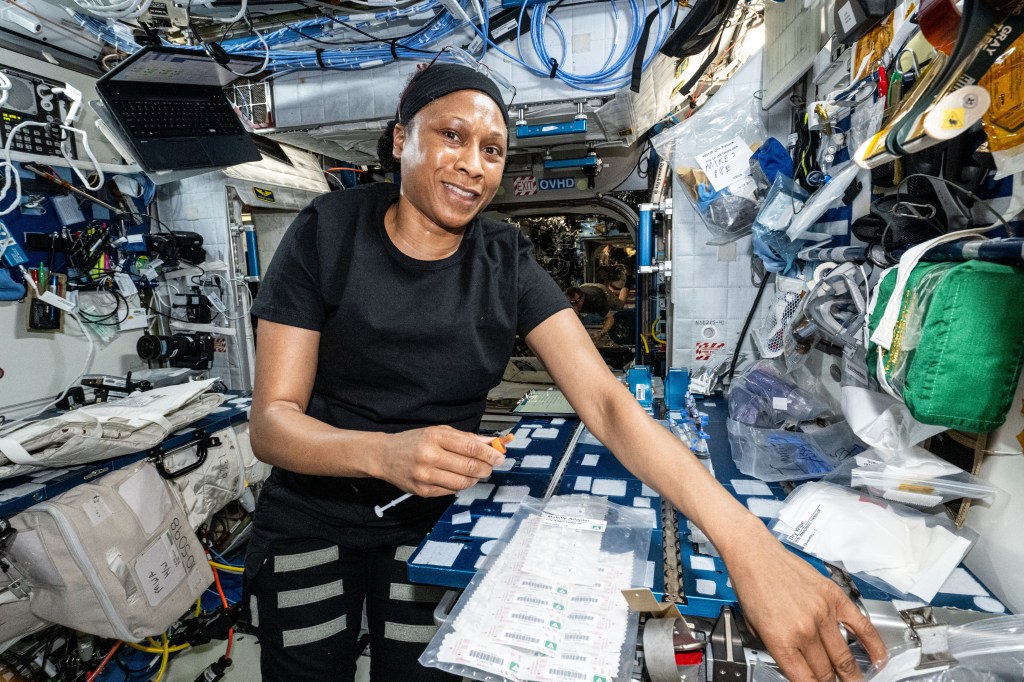 NASA astronaut Jeanette Epps has her left arm extended towards the camera. Epps holds a syringe in her right hand. Epps is looking at the camera with a slight smile, wearing a black t-shirt, black pants with Velcro strips, and a black headband.