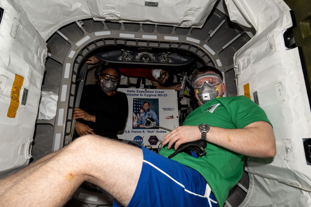 NASA astronauts (from left) Jeanette Epps and Matthew Dominick, both Expedition 71 Flight Engineers, are pictured wearing personal protective equipment (PPE) shortly after entering the Northrop Grumman Cygnus space freighter. Inside Cygnus is a poster of former NASA astronaut Francis R. "Dick" Scobee for whom the spacecraft is named after. The duo was wearing PPE after opening the hatch and entering the vehicle to protect against potential dust or debris that may have dislodged during Cygnus' launch and ascent to space.