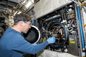 Barratt, wearing a long-sleeved blue shirt, glasses, a headlamp, and blue latex gloves, peers into the open round hatch of the combustion rack, using his right hand to pull out a device with two long metal protrusions and multiple cords connecting it to the rack.