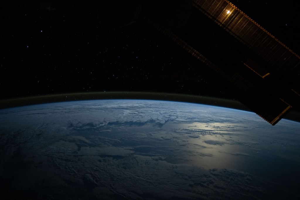 The Moon's glint beams off the Pacific Ocean as stars glitter in the background above the Earth's airglow. The Moon is obscured behind a solar array in this photograph from the International Space Station as it orbited 264 miles above.