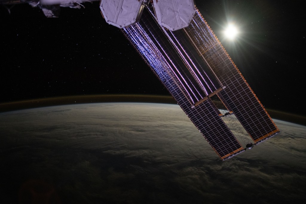 The Moon illuminates a cloud-covered Pacific Ocean as stars glitter in the background above the Earth's airglow. The International Space Station's solar arrays dominate the foreground as the orbital outpost soared 268 miles above.